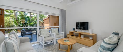 A modern living room with a large sliding glass door leading to an outdoor area, a flat-screen TV, a wooden coffee table, and a sofa with a patterned throw pillow.