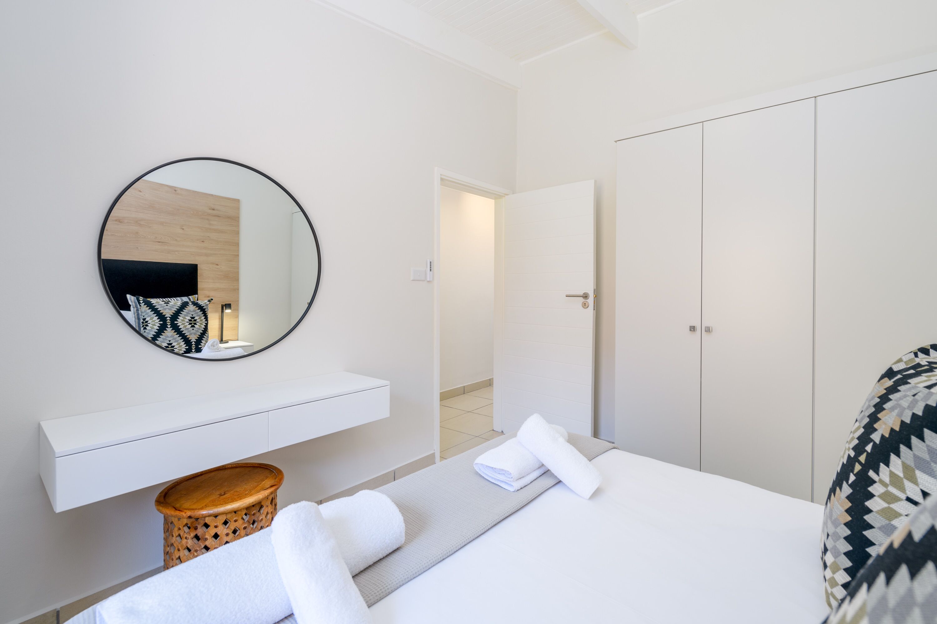 Quarto de hotel moderno com uma cama, um espelho redondo, uma mesinha de cabeceira, um guarda-roupa e um pequeno banquinho de madeira.