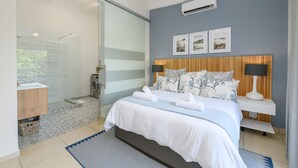 Una habitación de hotel moderna con una cama grande, cabina de ducha con mampara de vidrio, cabecero de madera y aire acondicionado de pared.