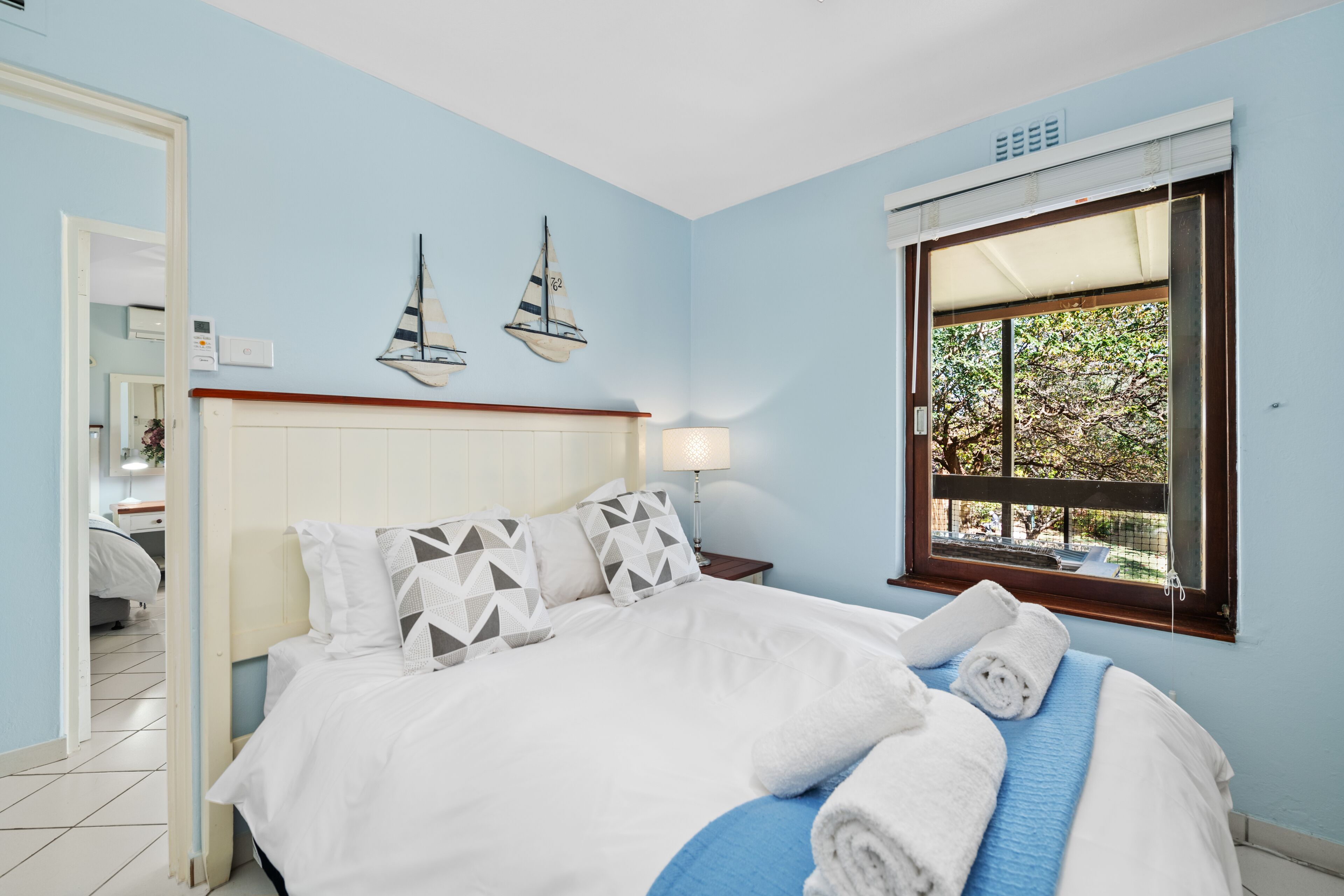Ein Schlafzimmer mit Bett, Kissen, einem Nachttisch, einer Lampe und einem Fenster mit Blick auf Grün.
