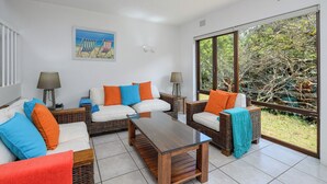 A modern living room with a white sofa, two side tables, a wooden coffee table, and a large window with a view of trees. - San Lameer Villa Rentals 3116 (Southbroom)