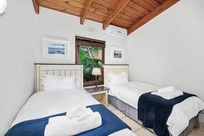 A hotel room with two single beds, a wooden ceiling, a window with a view, and an air conditioning unit.