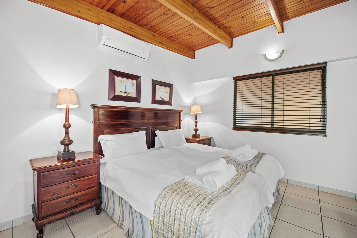 Une chambre avec un plafond en bois, un lit recouvert de draps blancs, deux tables de chevet avec des lampes et une fenêtre avec des stores.