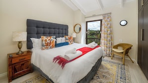 Une chambre avec un grand lit, une table de chevet, un fauteuil en osier et une vue sur l’extérieur.