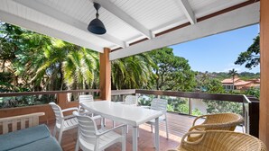 Une terrasse extérieure couverte, avec une table et des chaises blanches, des chaises en osier, et une vue sur des palmiers et des bâtiments en arrière-plan.