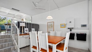 Une cuisine moderne avec des meubles blancs, une table à manger en bois et des chaises blanches. On aperçoit un escalier menant à un autre niveau, ainsi qu'un ventilateur de plafond.