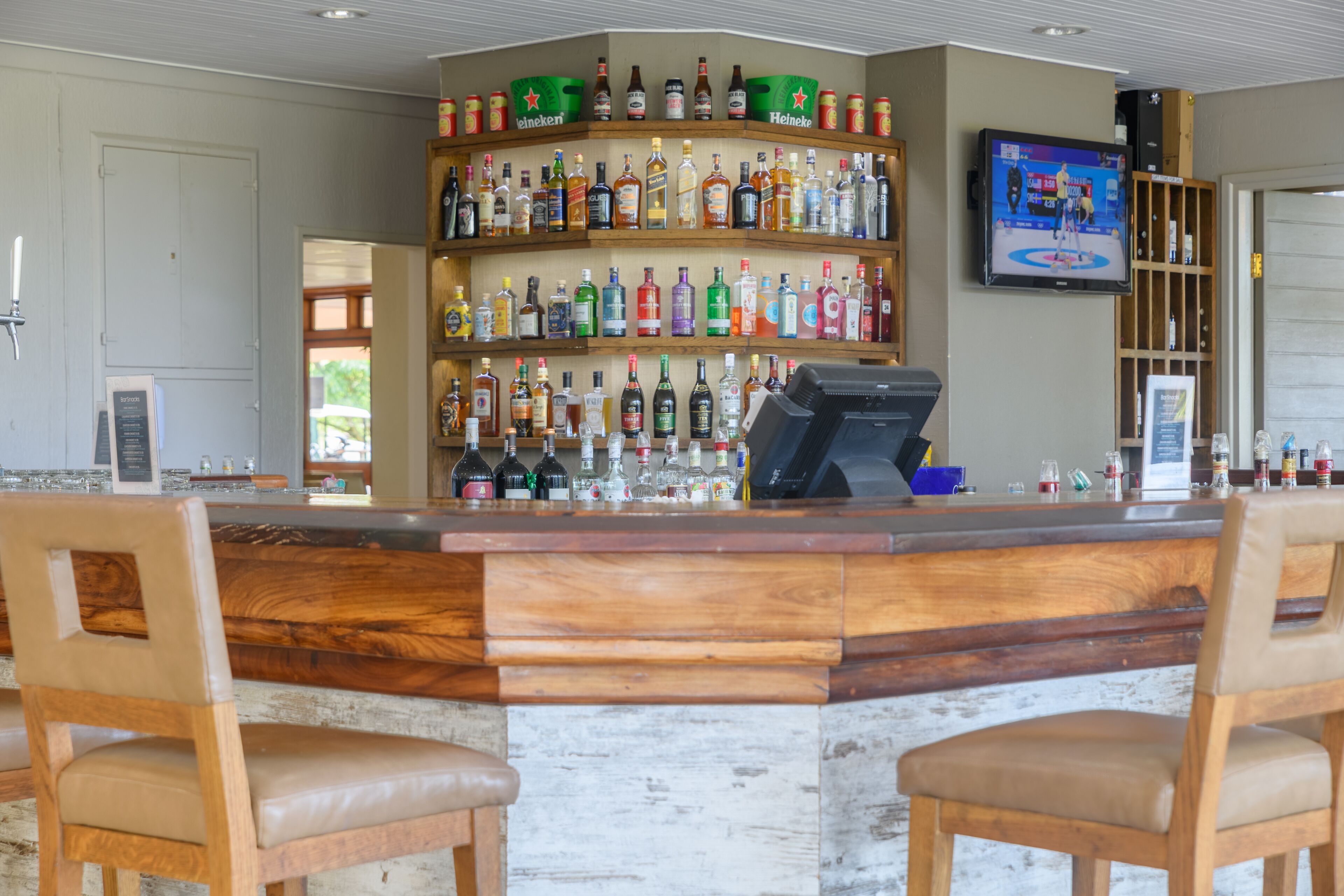Angolo bar con bancone in legno e due sgabelli, scaffali con diverse bottiglie di liquore, una televisione appesa al muro e una porta che dà sull'esterno.