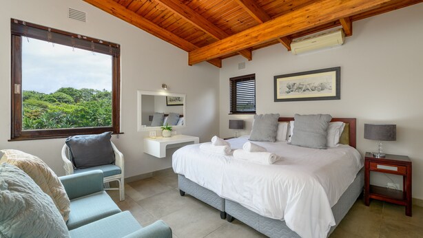 Quarto com cama grande, uma cadeira, mesa de cabeceira, espelho e janela com vista para área verde.