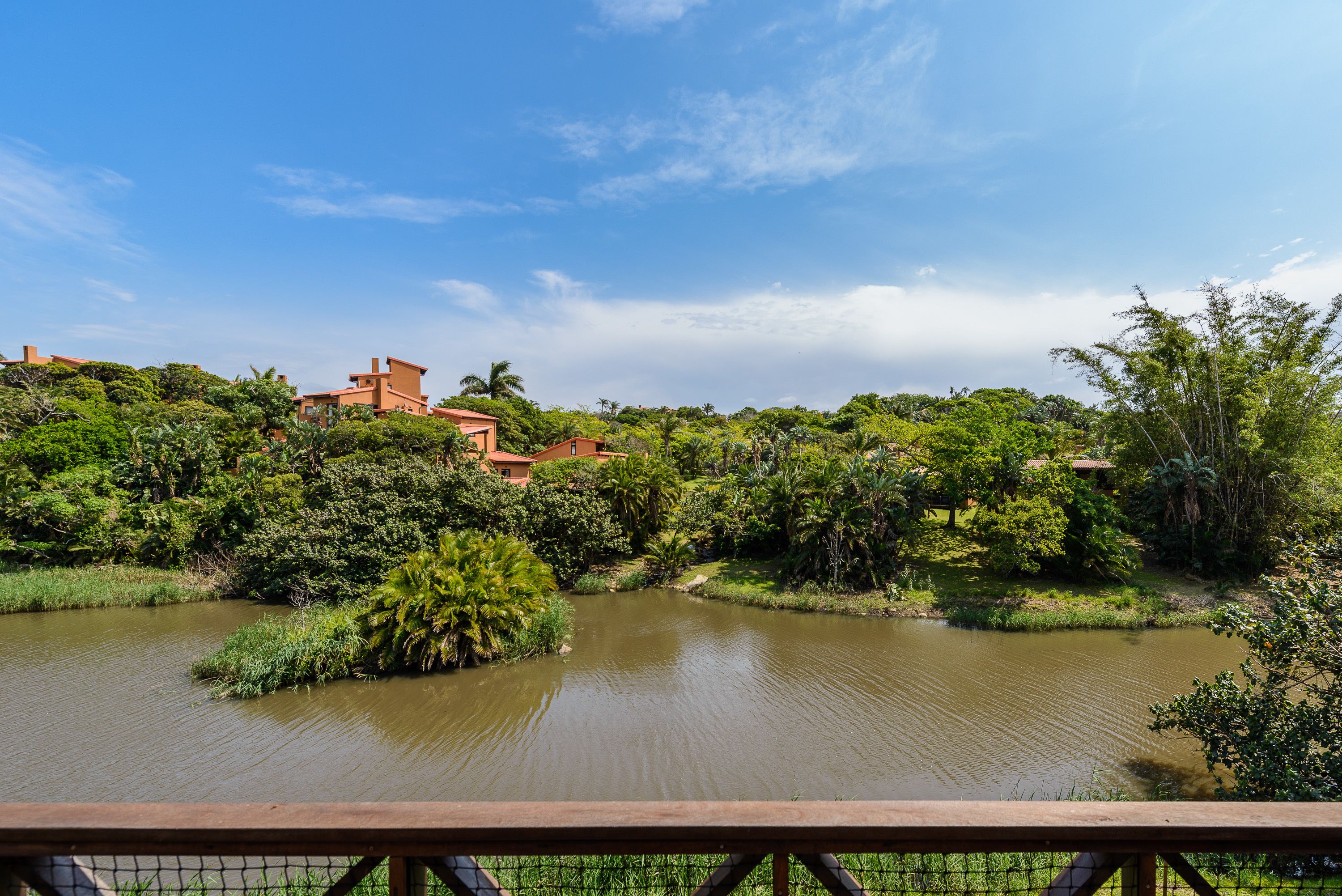 A river with a wooden bridge in the foreground, surrounded by lush greenery and buildings with red roofs.
