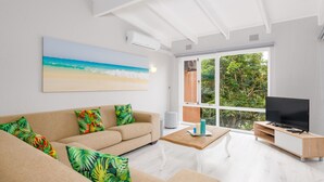 A modern living room with a beige sofa, colorful tropical pillows, a wooden coffee table, a flat-screen TV on a wooden stand, and a large window with a view of greenery. - San Lameer Villa Rentals 3108 (Southbroom)