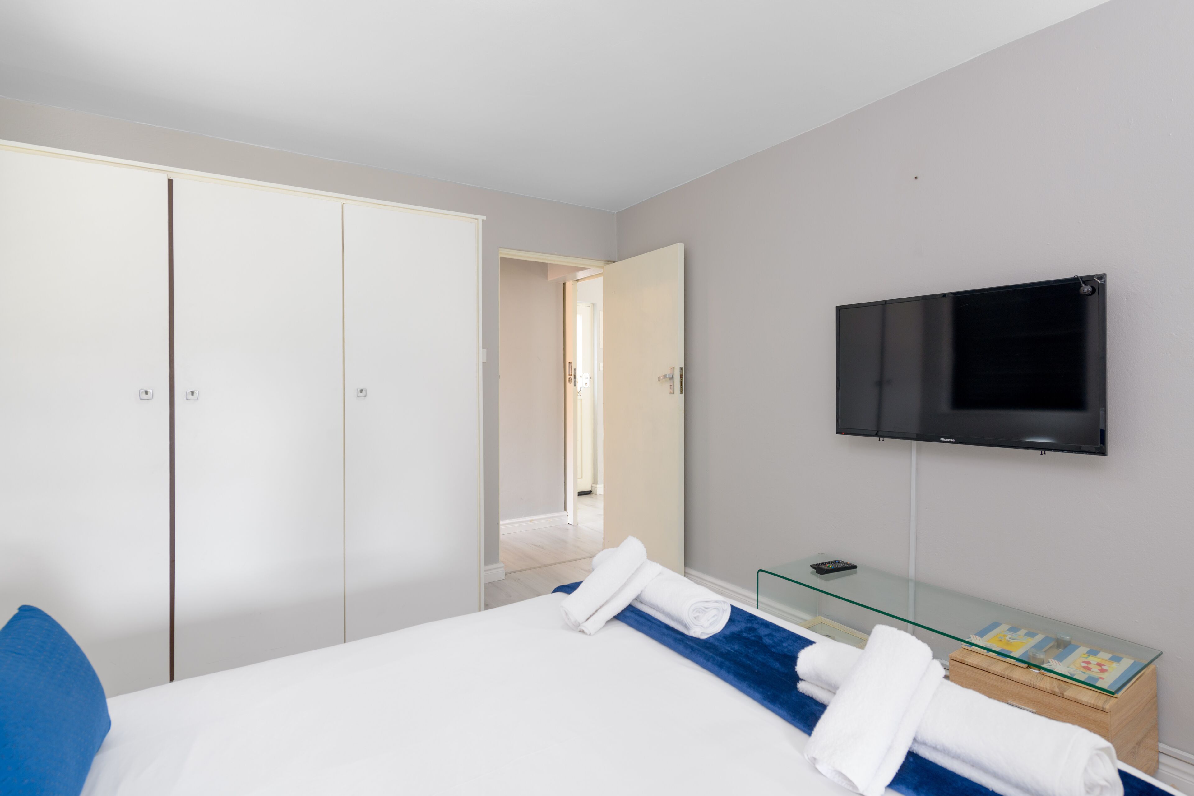 A hotel room with a bed, a TV mounted on the wall, a glass side table, and a wardrobe.