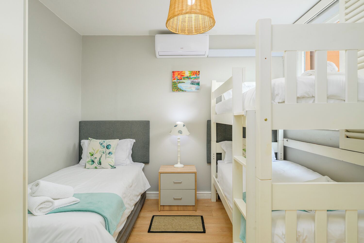 Quarto com uma cama de solteiro, mesa de cabeceira, luminária, beliches, ar condicionado e um quadro na parede.
