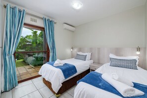 A hotel room with two beds, a window with curtains, an air conditioning unit, and a view of greenery outside. - San Lameer Villa Rentals 2529 (Southbroom)