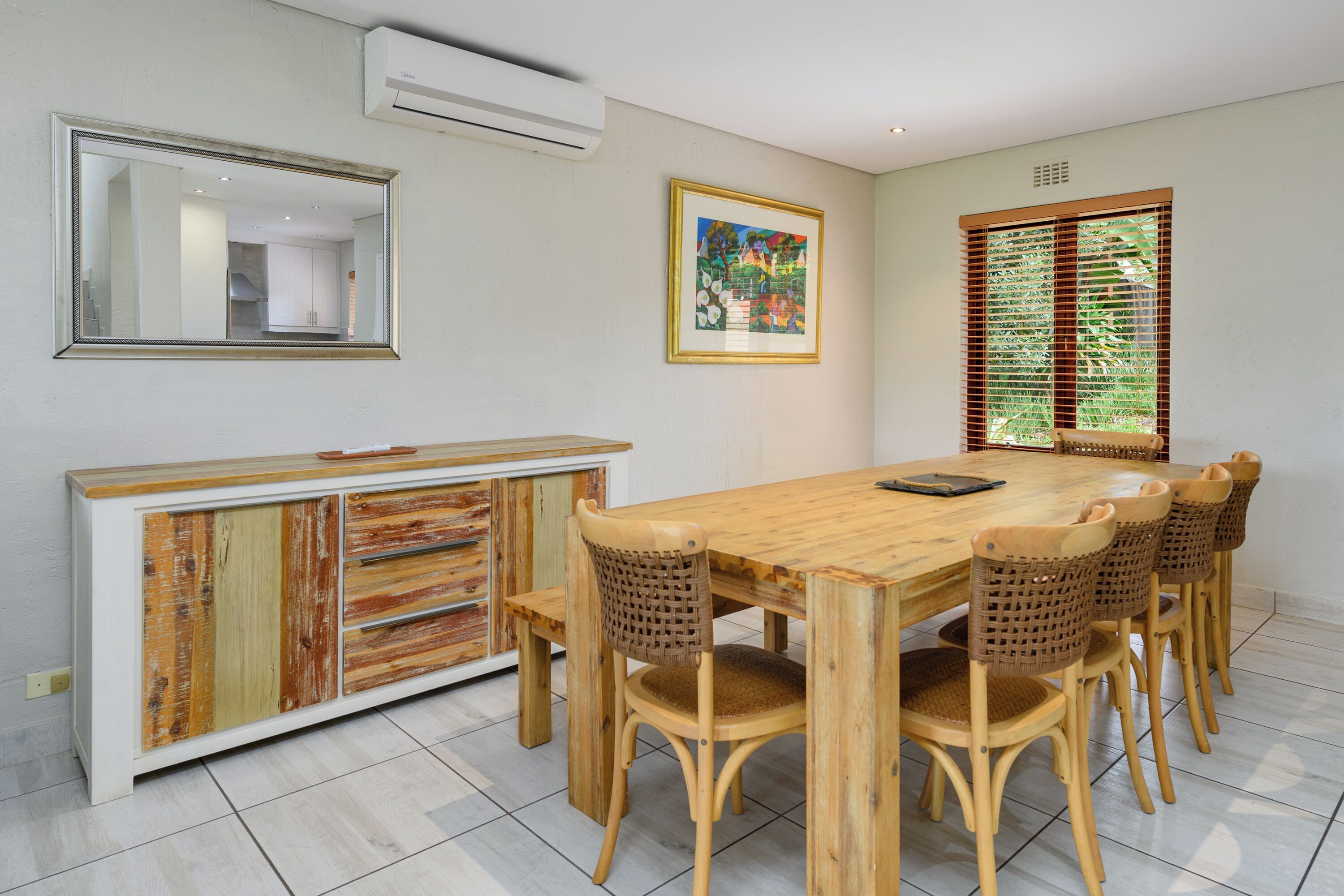 A dining area with a wooden table and chairs, a cabinet with a unique design, a framed picture on the wall, and a window with blinds.