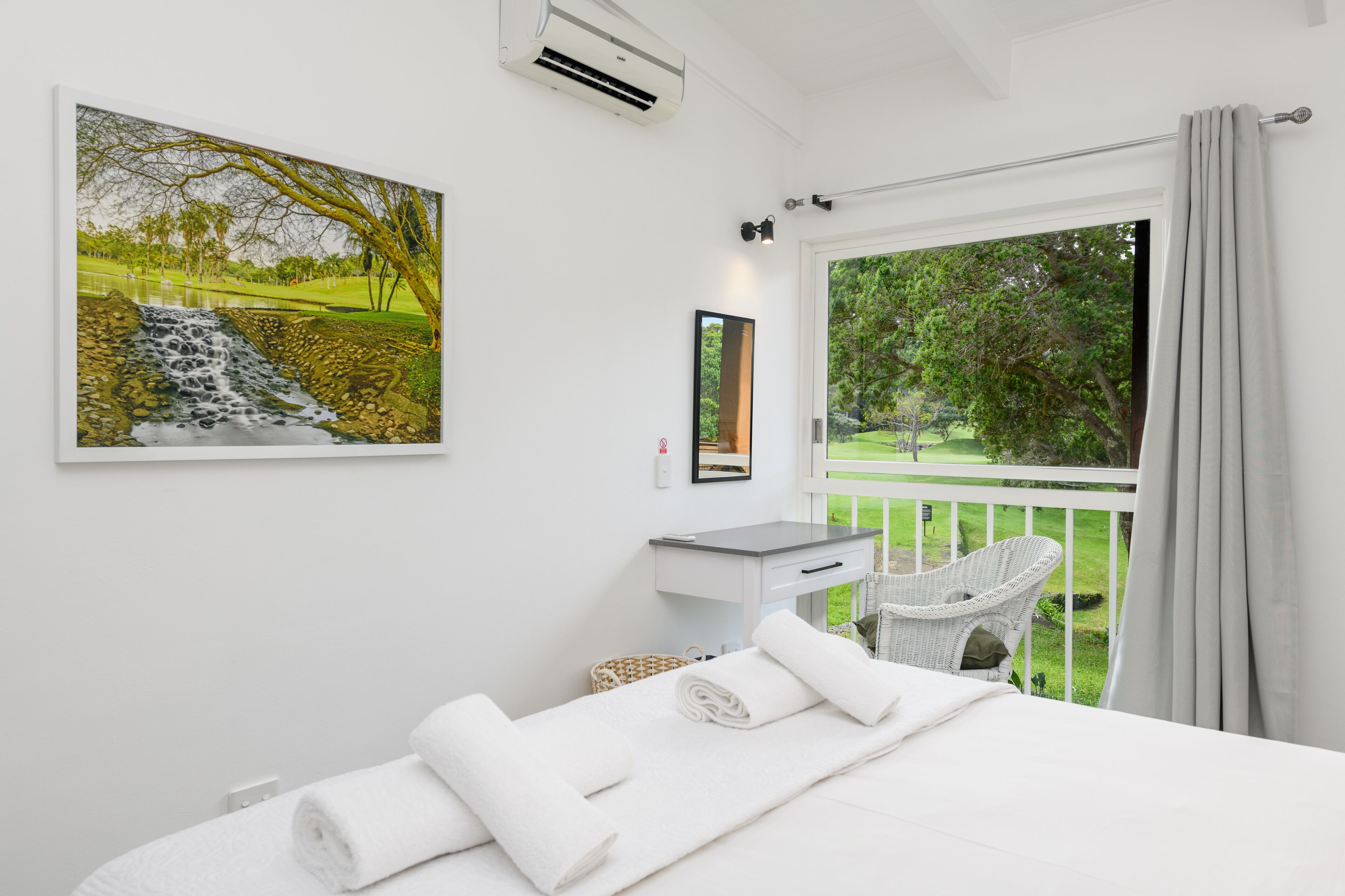 A bedroom with a bed, a white bedside table, a mirror, a window with a view of a garden, and a painting on the wall.