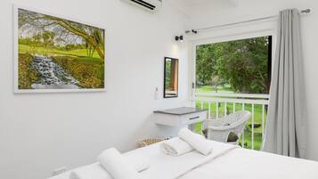 A bedroom with a bed, a white bedside table, a mirror, a window with a view of a garden, and a painting on the wall.