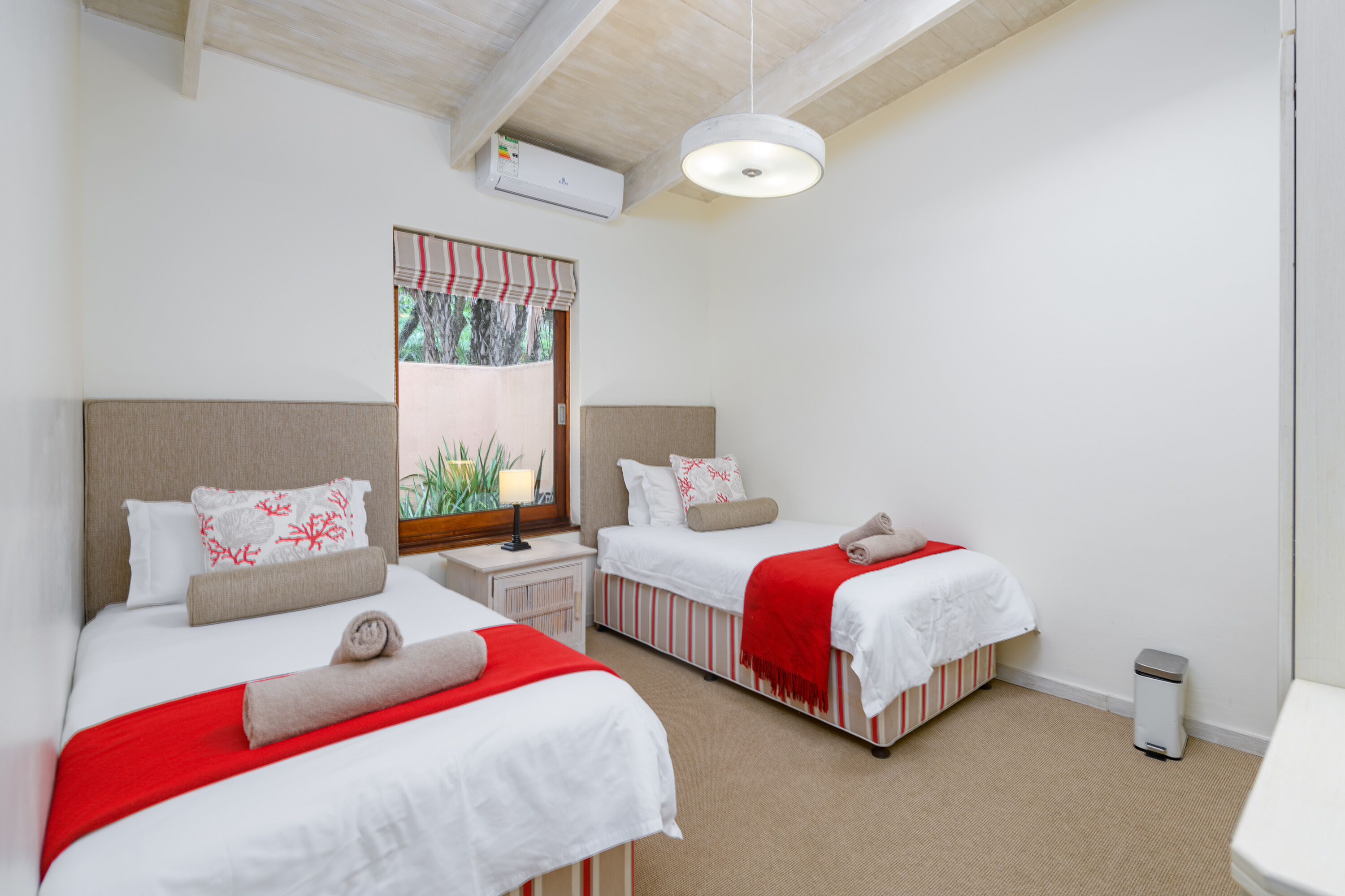 A hotel room with two beds, a window with a view of greenery, a ceiling fan, and a small bedside table with a lamp.