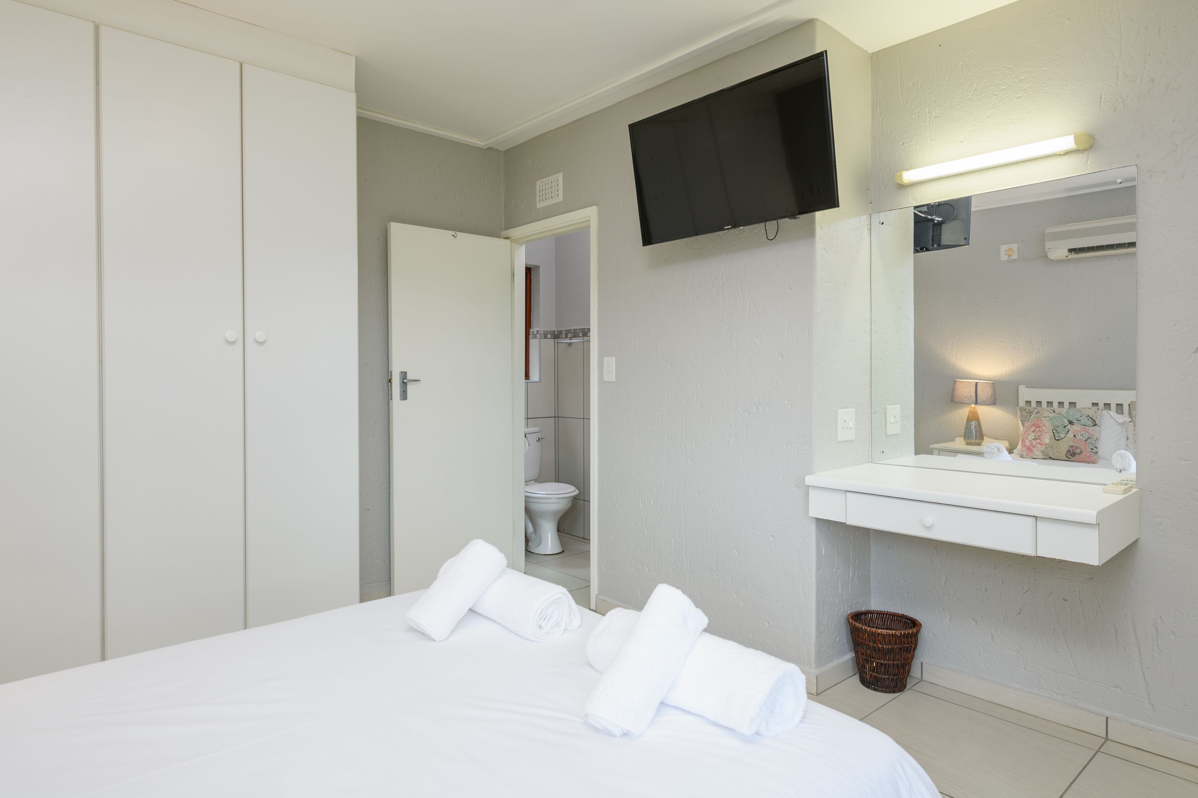 A hotel room with a bed, a TV mounted on the wall, a bathroom visible through an open door, and a white bedside table with a lamp.