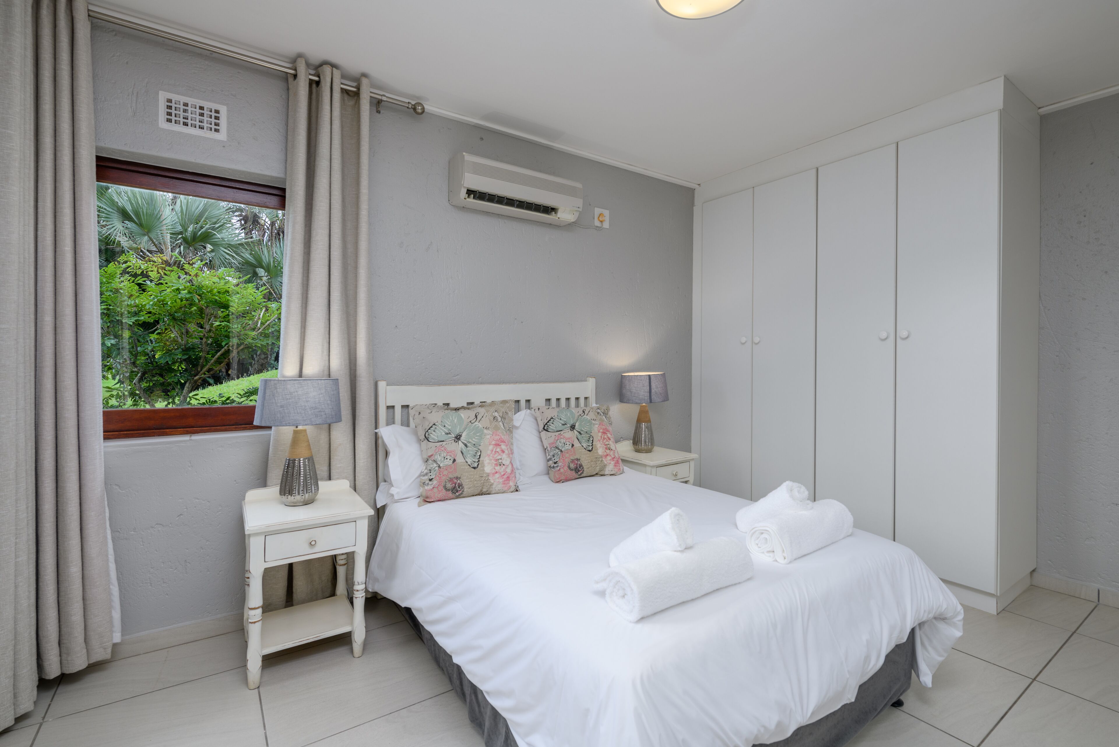 A bedroom with a bed, a nightstand, a lamp, a window with curtains, an air conditioning unit, and a wardrobe.