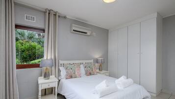 A bedroom with a bed, a nightstand, a lamp, a window with curtains, an air conditioning unit, and a wardrobe.