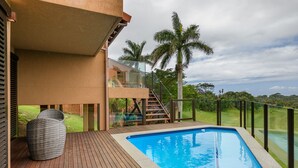 A modern outdoor pool area with a wooden deck, a glass balustrade, and a view of a golf course. - San Lameer Villa Rentals 14109 (Southbroom)