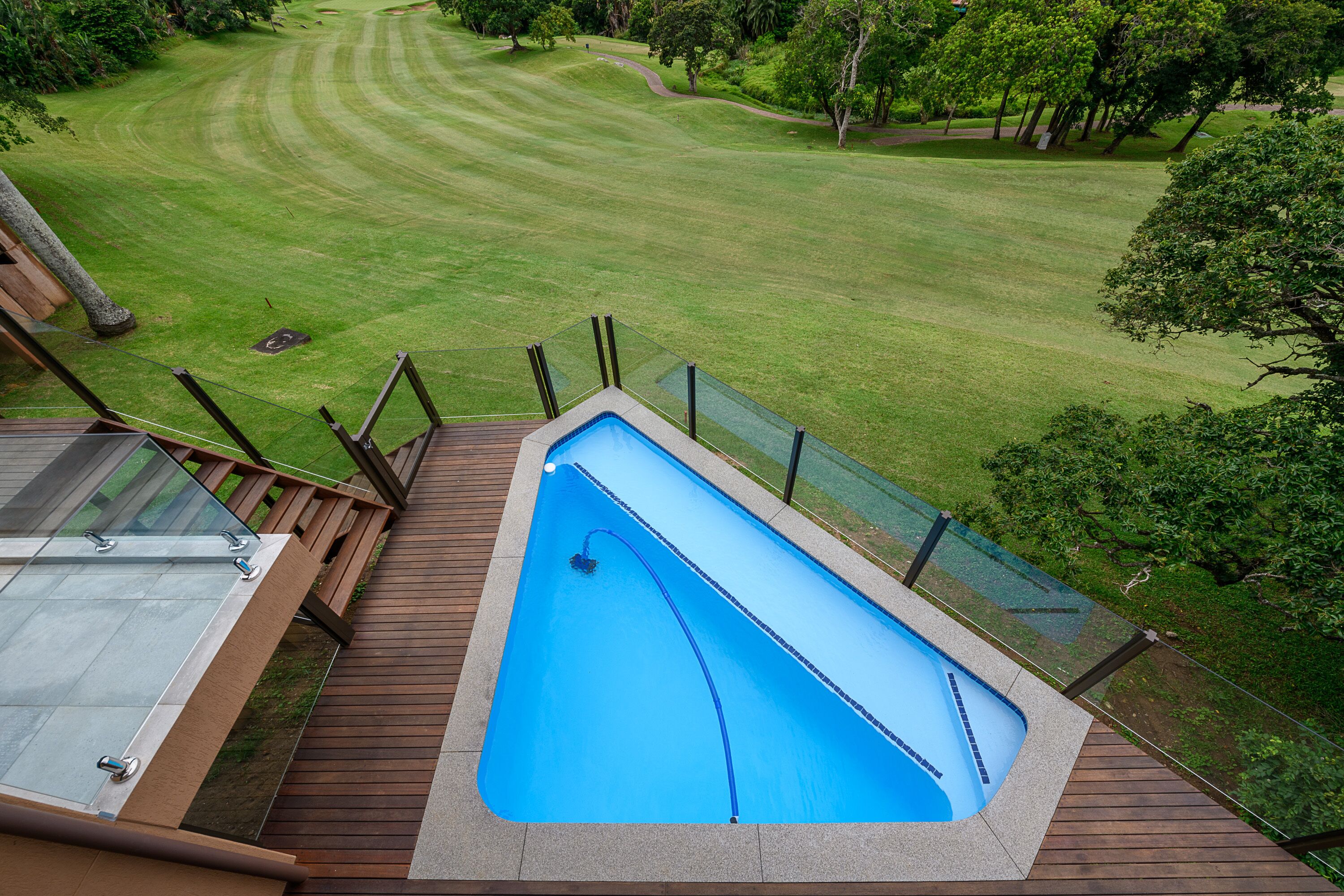 Une piscine dotée d’une terrasse en bois et de rampes de protection en verre, entourée d’une pelouse bien entretenue et d’arbres.
