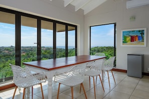 A dining area with a wooden table and white chairs, large windows with a view of greenery, and a painting on the wall.