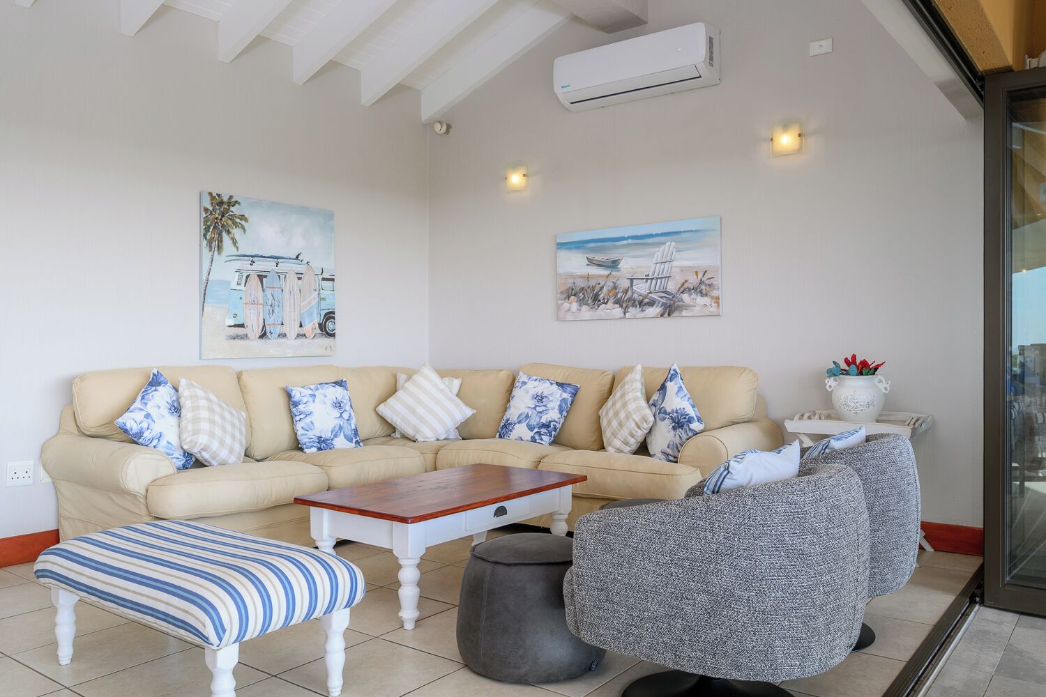 A living room with a beige sofa, striped bench, and grey armchair, with two wall-mounted air conditioners and paintings.