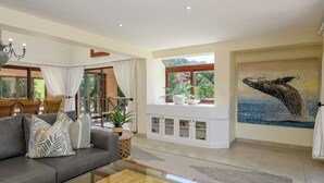 A spacious living room with a grey sofa, a glass coffee table, and a white cabinet with decorative items. There is a large window with a view of greenery and a whale painting on the wall. - San Lameer Villa Rentals 14014 (Southbroom)