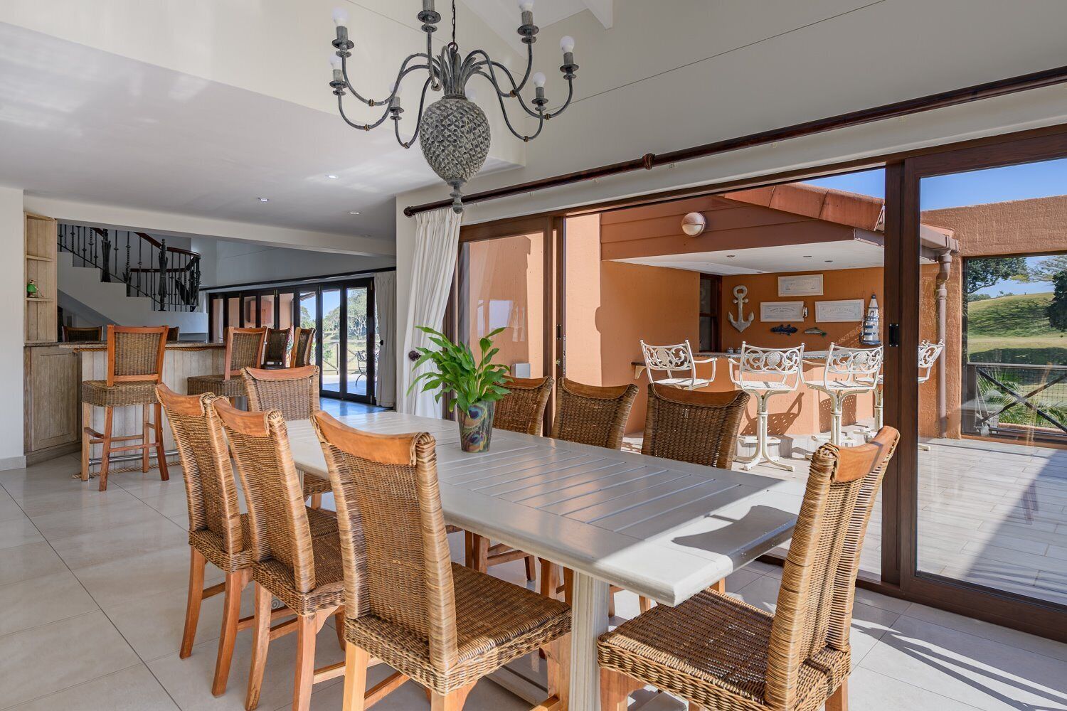 A spacious dining area with a large table and wicker chairs, a chandelier, and a view of the outdoors.