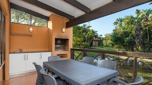 An outdoor kitchen with a table and chairs, a grill, and a view of a garden and pond. - San Lameer Villa Rentals 1902 (Southbroom)