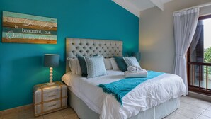 A bedroom with a large bed, a nightstand, a lamp, a suitcase, and a window with curtains. - San Lameer Villa Rentals 1929 (Southbroom)