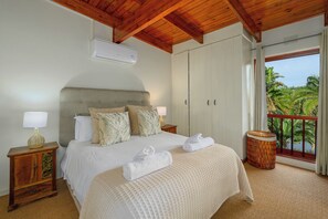 A bedroom with a bed, two nightstands, an air conditioner, a window with a view, and a balcony with a wicker stool. - San Lameer Villa Rentals 3506 (Southbroom)