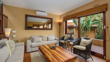 A living room with a sofa, a wooden coffee table, wicker chairs, a large mirror, and a view of greenery through a glass door.