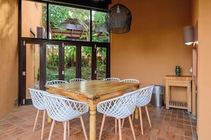 A dining area with a wooden table and white lattice chairs, a hanging light fixture, and a view of greenery through large glass doors.