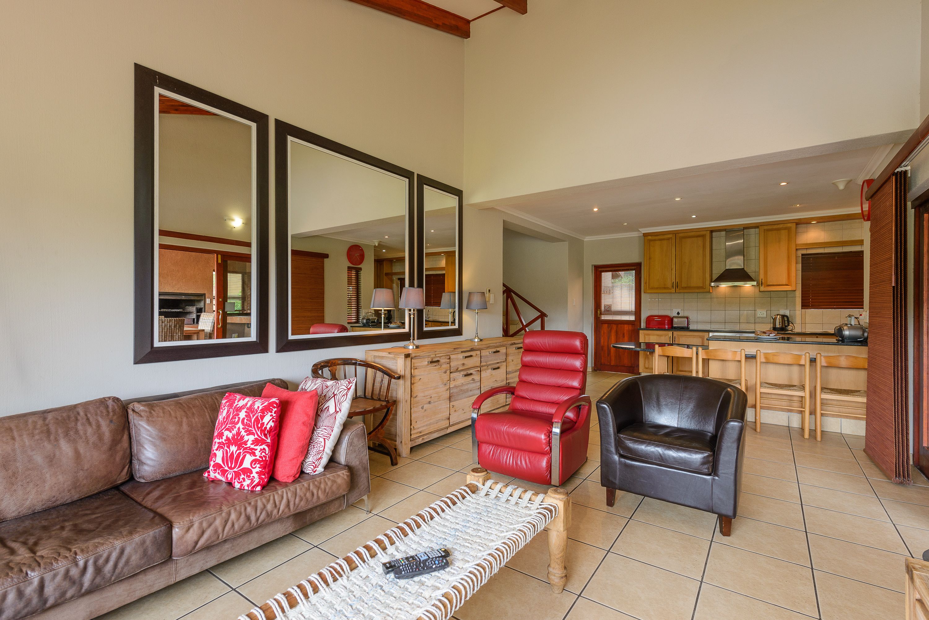 Amplia sala de estar con un sofá de cuero marrón, un sillón de cuero rojo y una mesa de centro de madera. Un espejo grande cubre la pared, reflejando la cocina con armarios de madera y un salpicadero de azulejos.