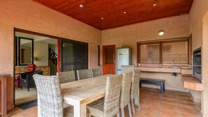 Une terrasse couverte avec un plafond en bois, une table à manger avec des chaises en osier, un réfrigérateur et une fenêtre avec des stores.