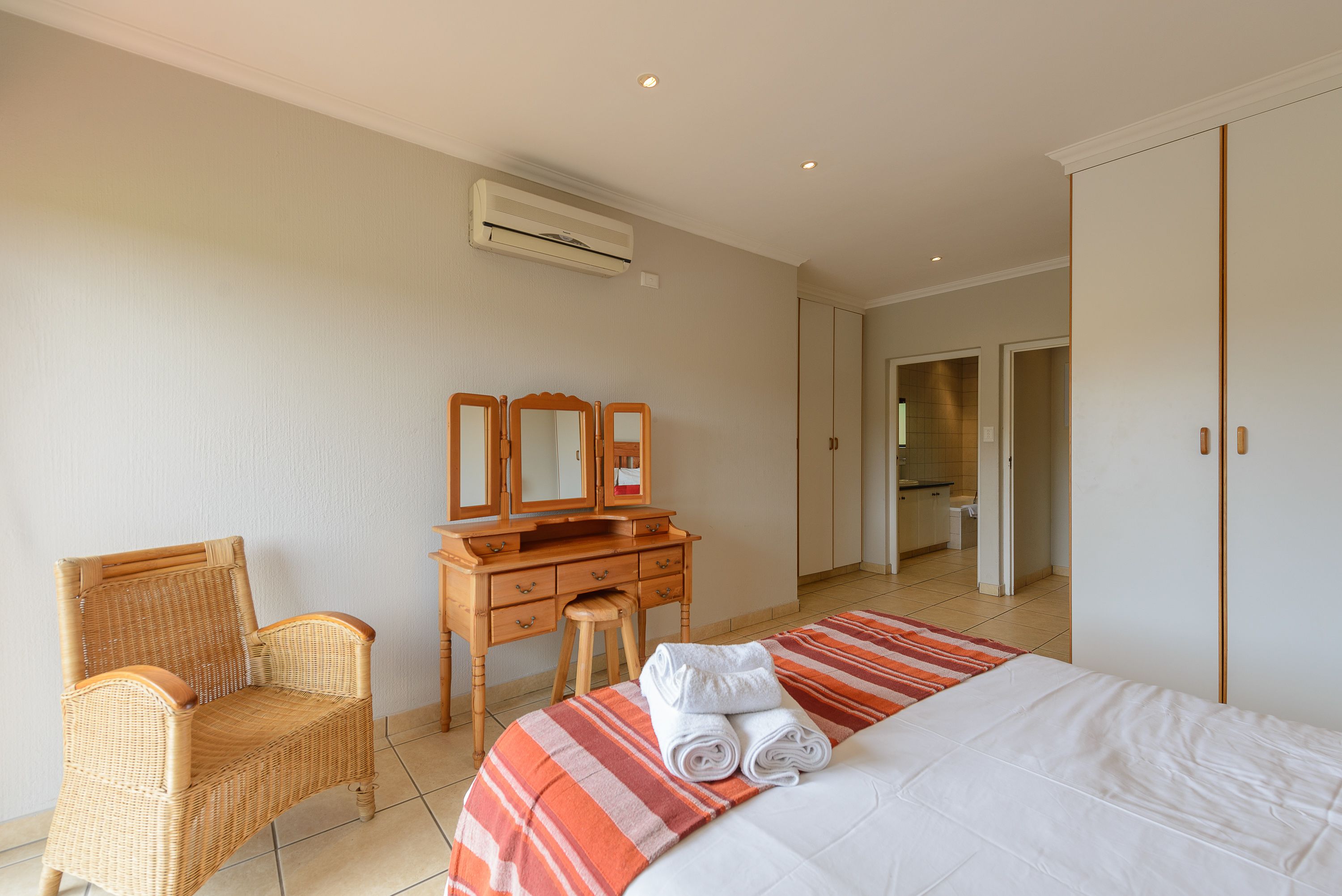 Quarto de hotel com cama, cómoda de madeira com espelho, cadeira de vime e ar condicionado na parede.