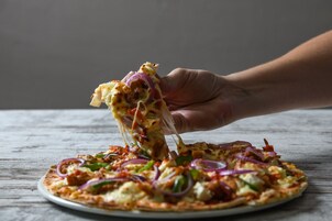 Una mano sosteniendo una porción de pizza con ingredientes, sobre una superficie de madera.