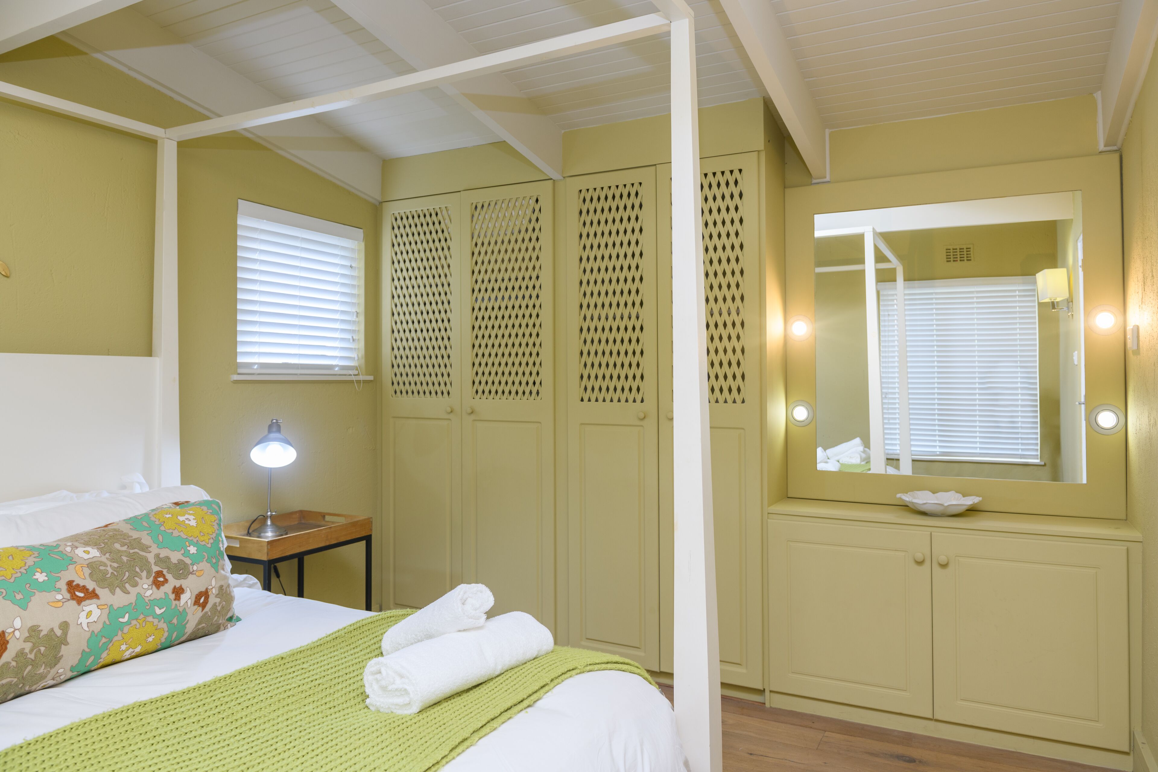 A bedroom with a bed, a bedside lamp, a green blanket, a pillow, a mirror, and a wooden nightstand.