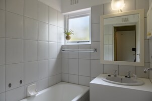 Un baño con azulejos blancos, una ventana con una planta, una bañera y un lavabo con espejo.