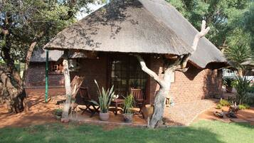 A rustic thatched-roof hut with a brick exterior, surrounded by potted plants and a small outdoor seating area.
