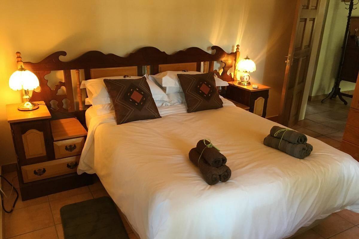 A neatly made bed with white linens, two pillows, and two rolled towels on the bed. There are two bedside tables with lamps and a small drawer unit.