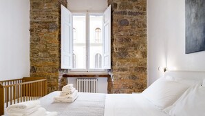 A bedroom with a stone wall, a wooden crib, a bed with white linens, and a view through a window with open shutters. - Luxury 2 Bedroom Apartment with Hot Tub - Florence City Center (Florence)