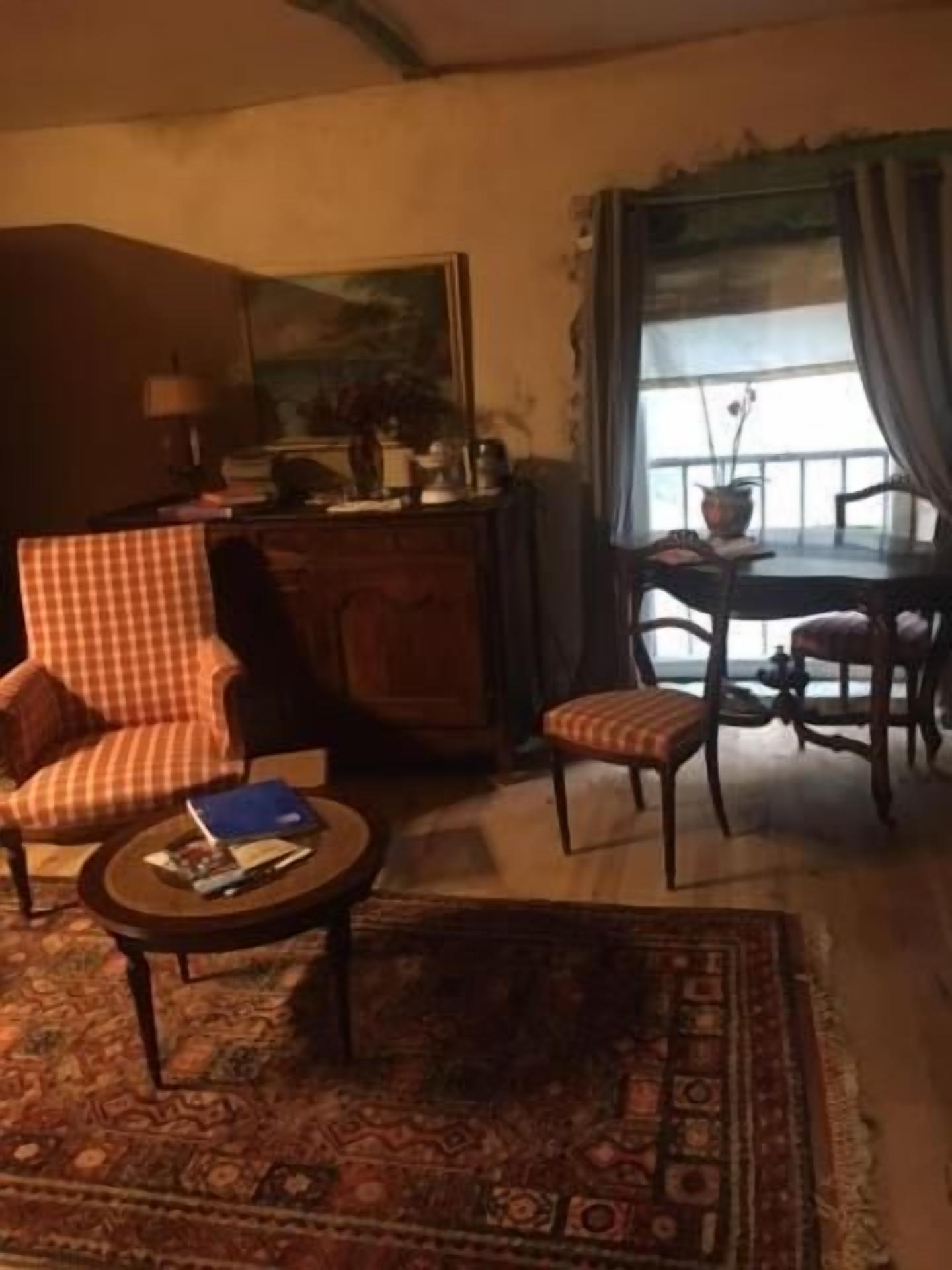 Een woonkamer met een geruite fauteuil, een klein rond tafeltje met een boek, een kamerkleed met dessin, een eettafel met stoelen en een uitzicht door een raam met gordijnen.