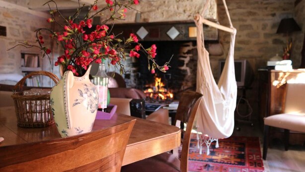 A rustic interior with a wooden table, a hanging chair, and a fireplace in the background. - Le Moulin à Huile (Vailhourles)
