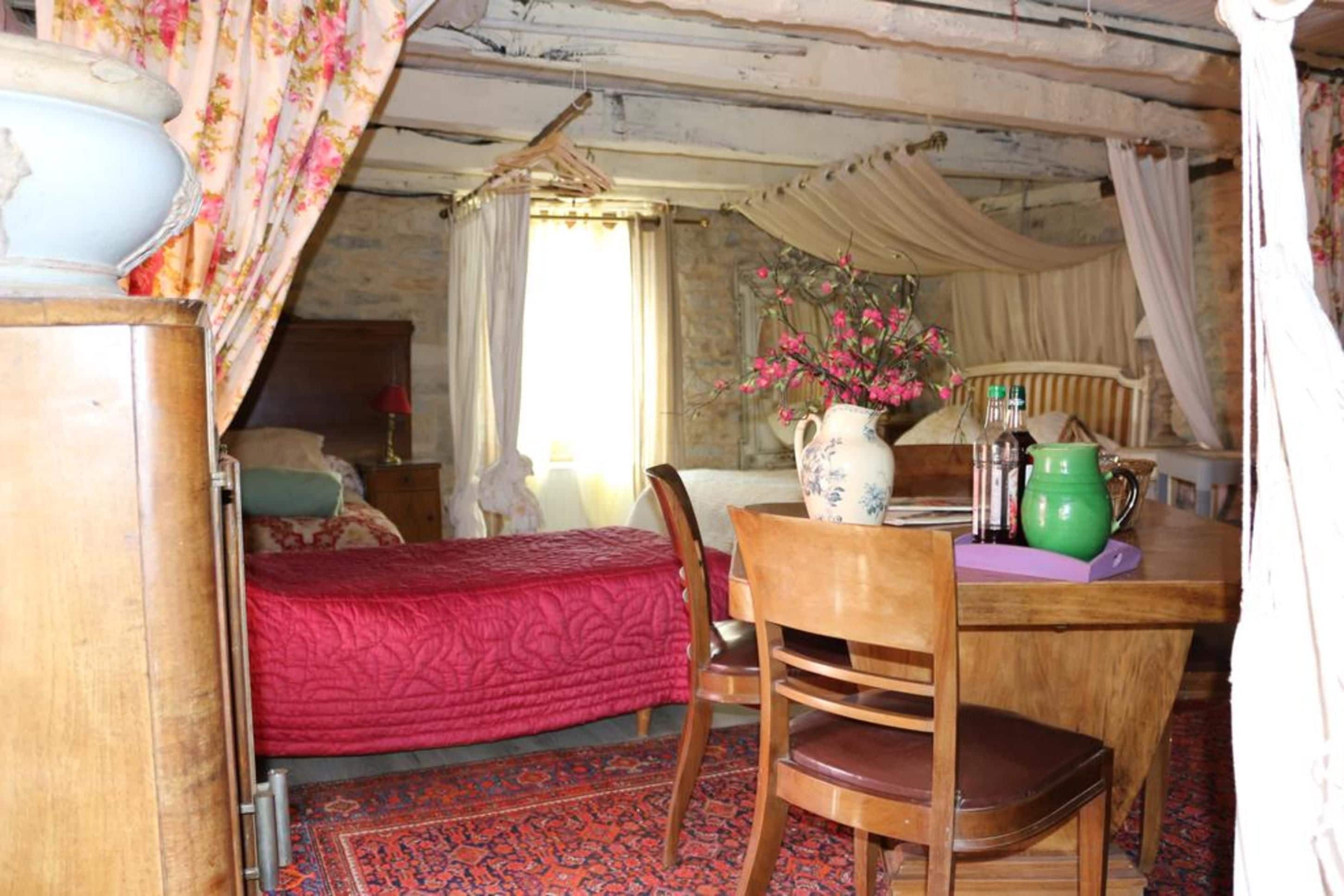 Une chambre confortable avec un lit, une chaise en bois, une table ornée d’un vase de fleurs et une fenêtre avec des rideaux.