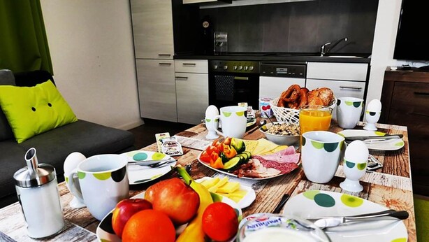 A breakfast table set with plates of fruit, eggs, toast, and juice in a modern kitchen with a sofa and green curtains in the background. - Speedys Gästehaus am Brünnchen (Herresbach)