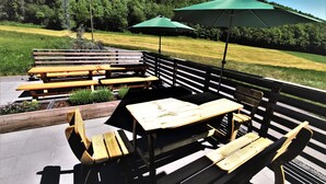 An outdoor patio with wooden benches and tables, green umbrellas, and a view of a grassy field and forest. - Speedys Gästehaus am Brünnchen (Herresbach)