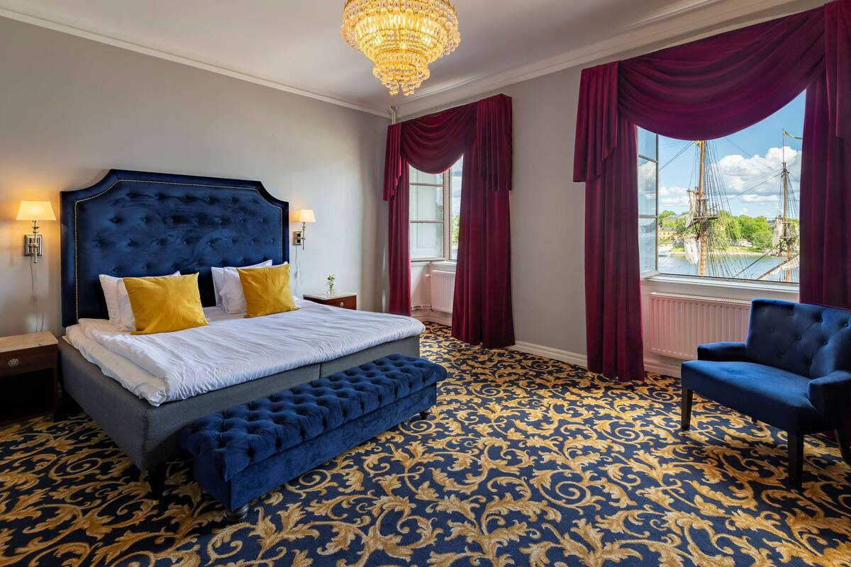A bedroom with a large bed, a blue upholstered bench, a blue armchair, and a view of a river with ships.
